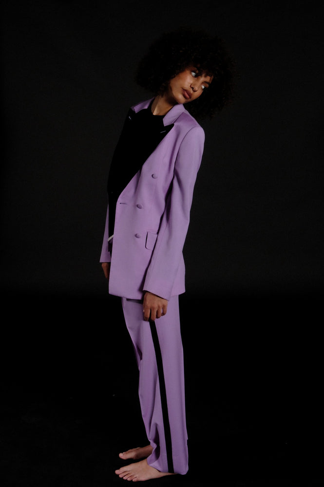 
                  
                    A woman is wearing a lilac double breasted tuxedo with black lapels covered in velvet
                  
                